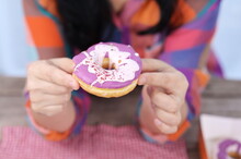 A Woman Holds A Delicious Donut Coated In Purple And White Sugar, Topped With Crushed Red Flower Petals. A Woman In A Bright Shirt Holds A Donut And Thrusts It Forward To Invite People To Eat.
