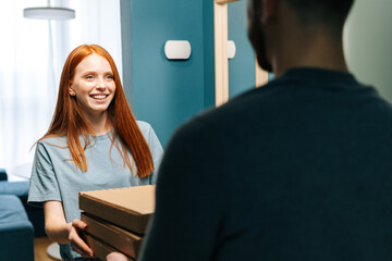 Cheerful young woman receiving paper boxes with hot pizza from unrecognizable delivery man on doorstep at home. Back view of courier male delivering boxes with food to female client at apartment.