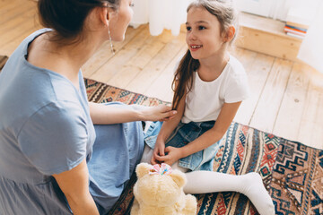 Pretty happy stylish Caucasian mother play with her cute little daughter on floor on fashionable carpet at home, touching her hand gently, talking, giving advice, child listening with interest