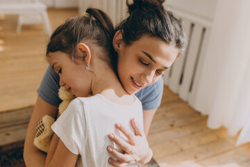 Love and care in family. Strong bonds. Human relationships. Mother and daughter. Young charming Caucasian female embracing gently her child, sitting on floor in living-room, girl holding teddy bear