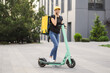 Delivery woman using smartphone while standing in the city with electric push scooter. Adult delivery woman in black t-shirt and jeans delivering online order to client.