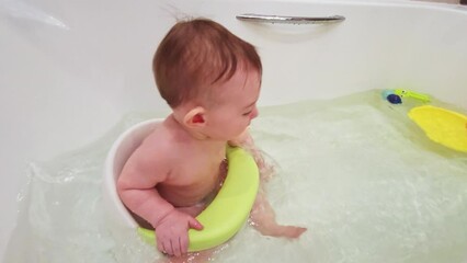 happy toddler baby boy bathes in a white bath while sitting on a chair. child playing with water in 