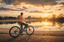 Woman Riding A Mountain Bike Near Lake At Sunset In Summer. Colorful Landscape With Sporty Girl Riding A Bicycle, Beach, Beautiful Sky Reflected In Water In Park In Spring. Sport And Travel. Biking