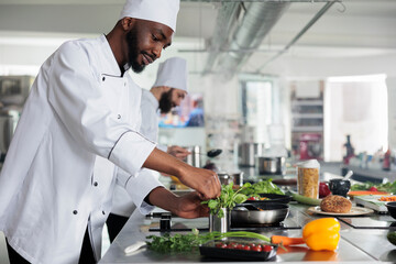 Wall Mural - African american professional head chef picking fresh green herbs to improve gourmet dish taste. Male cook wearing cooking uniform preparing organic meal while in restaurant kitchen.