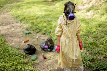 Woman in gas mask and protective clothes stands with garbage bags collecting scattered plastic in the woods. Problem of environmental pollution. Safe the world concept