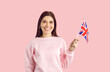 Portrait of happy smiling young woman with flag of UK. Pretty British girl with cheerful face expression standing isolated on pink background and holding Union Jack. Learning English language concept