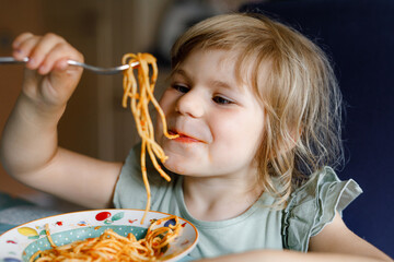 Adorable toddler girl eat pasta spaghetti with tomato bolognese with minced meat. Happy preschool child eating fresh cooked healthy meal with noodles and vegetables at home, indoors.