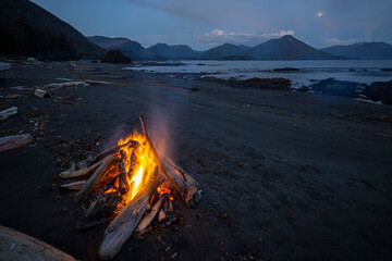 Campfire on calm lake shore with mountains in the background