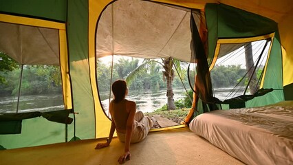 Wall Mural - Asian woman relaxing inside a tent and taking the riverside view in the sunset on vacation