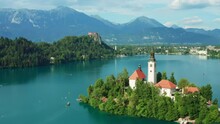 Fly Around Pilgrimage Church Of Assumption Of Mary Built On Island Covered With Lush Green Trees. Bled Lake With Blue Water Among Forestry Mountains. 