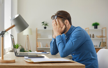 Young businessman receives letter with bad news. Upset tired man sitting at working desk with laptop and correspondence and hiding his face in his hands. Stress and business problems concept