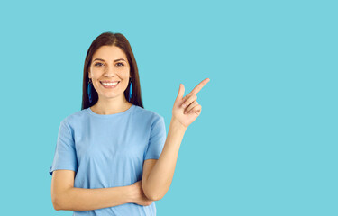 Happy woman standing and showing something on blue copyspace background. Attractive brunette lady in T shirt and earrings points with index finger at contact number or asks to follow social media link