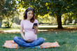 A pregnant woman in headphones listens to music and uses the phone in the park on a summer day. Place for text.