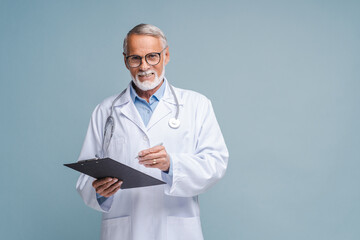 Wall Mural - Mature physician therapist writing down treatment plan, writes a prescription. Senior doctor in white medical gown with stethoscope on shoulders taking notes standing with clipboard isolated on blue