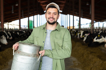 Closeup portrait of cheerful young bearded dairy farm owner carrying aluminum can of milk in cowshed against background of cows in stall