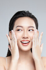 Portrait of cheerful laughing Asian woman applying foam for washing on her face with attractive appearance. Skincare spa relax concept. Isolated on grey background