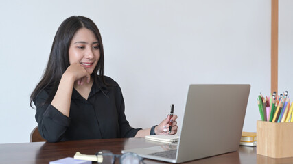 Wall Mural - Happy young asian woman looking at laptop computer screen and making notes on notebook.