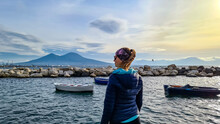 A Tourist Woman Enjoying The Panoramic View From The Harbor Of The City Of Naples, Campania, Italy, Europe. Bathing In The Sun. Small Fishermen Boat Floating In The Port. Walking Along The Sea Coast