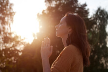 Peaceful Woman Holds Her Palms In Front Her In Prayer Against Backdrop Trees On The Lake In The Rays Of The Setting Sun. Meditating Or Praying. Caucasian Woman Believes In A Higher Power