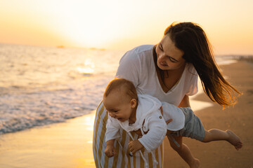 Wall Mural - happy mother's day. Beautiful mother and baby play on the beach. Mum and her Child together enjoying sunset. Loving single mother hugs cute little son.