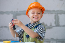 Kid Boy Twists Bolt With Screwdriver. Child Repairman With Repair Tool. Child In Helmet And Boilersuit On Construction Site. Little Worker Engineer.