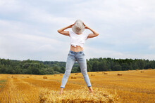 Girl In Straw Hat Stands On A Haystack On A Bale In The Agricultural Field After Harvesting