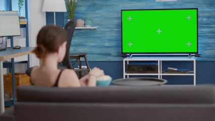 Wall Mural - Static tripod shot of young woman watching movie on tv with green screen and eating popcorn while sitting on couch. Girl looking at favourite television show on chroma key display in home living room.