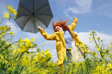 A woman with a blue shirt and a yellow raincoat jumping in the field of yellow rapeseed and having fun on a sunny summer day