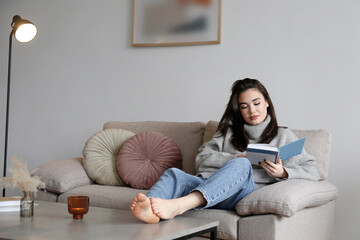 Young beautiful woman wearing grey turtleneck reading a book at home. Brunette female in a sweater sitting on beige textile couch in her lofty apartment. Background, copy space, close up.