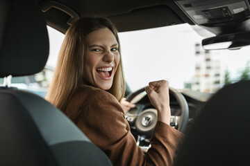 Excited young woman celebrating successful car purchase, gesturing YES while sitting inside auto salon at dealership