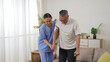 unwell asian elderly male with knee pain standing up slowly with a stick from the sofa at home. female nursing aide giving support while the man groans painfully