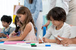 © Siam Stock - Selective focus of a little cute Caucasian boy sitting at a small table, drawing intentionally together with multi-ethnic friends, with teachers standing watching them in the back at kindergarten.