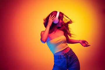 Portrait of young happy girl listening to music in headphones and dancing isolated over gradient red yellow background in neon