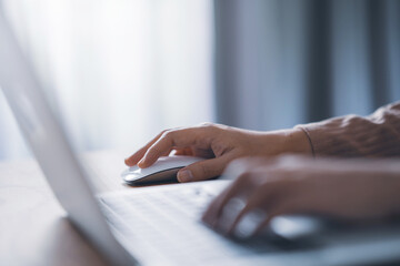 Close up finger of woman using modern white wireless mouse while working on laptop computer on wooden table at home.