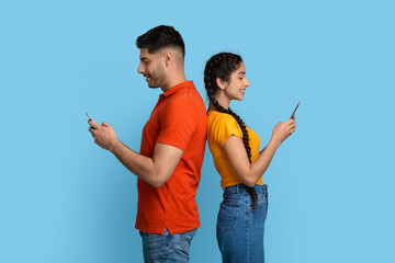 Cheerful middle eastern couple standing back to back and using smartphones