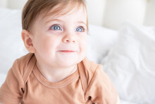 Portrait Of Cute Baby Girl 10 Months With Blue Eyes In Brown Bodysuit On White Bedding On Bed