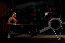 Serious Young Lady Exercising On Rowing Machine In Dark Gym