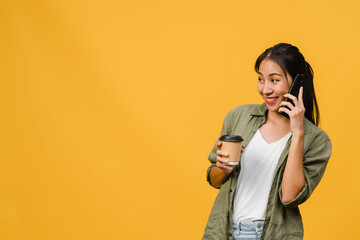 Wall Mural - Young Asia lady talk by phone and hold coffee cup with positive expression, smile broadly, dressed in casual cloth feeling happiness and stand isolated on yellow background. Facial expression concept.