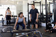 Strong Woman Looking Ahead Squatting While Doing A Deadlift With A Trap Bar While Her Trainer Looks On.