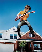 Teenage Boy With Guitar Jumping In Front Of Home, Striking A Pose Mid Air