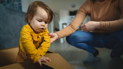 Wall Mural - One small Caucasian baby girl playing by her mother in the cardboard box at home sitting in the box real people family and growing up concept copy space