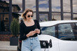 © Roman - Young beautiful woman traveling by electric car having stop at charging station standing plugging cable browsing internet on smartphone smiling joyful while charing