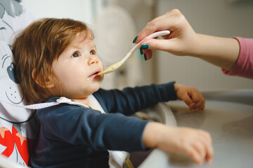 One small caucasian baby feeding while sitting in the chair hand of unknown mother giving food to small seven months old infant with plastic spoon in room at home real people care concept copy space