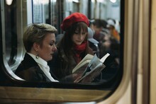 Young Caucasian Women Sitting In Subway Carriage In Saint Petersburg, Russia. One Is Reading A Book, Other Looking At Smartphone Image With Selective Focus, Toning And Noise Effect