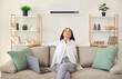 Woman enjoying modern AC system in her home. Happy relaxed middle aged lady breathing in fresh air while sitting on comfortable sofa with laptop computer in living room with air conditioner on wall
