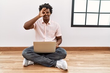 Poster - African man with curly hair using laptop sitting on the floor doing ok gesture with hand smiling, eye looking through fingers with happy face.