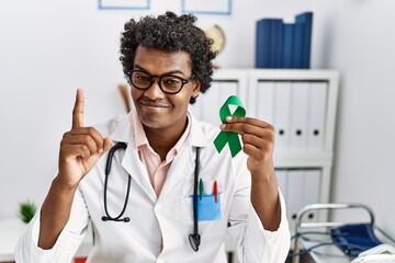 Sticker - African doctor man holding support green ribbon surprised with an idea or question pointing finger with happy face, number one