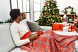 Young african american man sitting on the sofa drinking coffee by christmas tree looking to side, relax profile pose with natural face and confident smile.