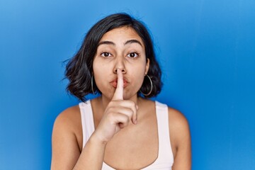 Wall Mural - Young hispanic woman standing over blue background asking to be quiet with finger on lips. silence and secret concept.