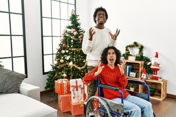 Poster - Young interracial couple with woman sitting on wheelchair by christmas tree crazy and mad shouting and yelling with aggressive expression and arms raised. frustration concept.